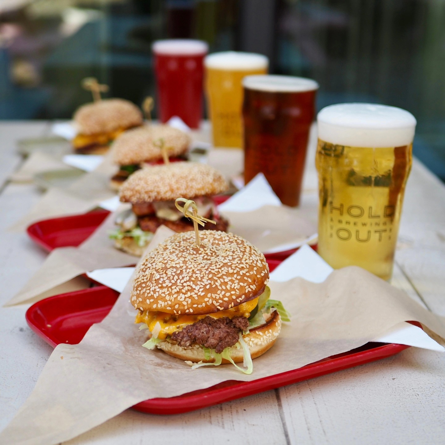 Four burgers on red trays and four beers on a white table.
