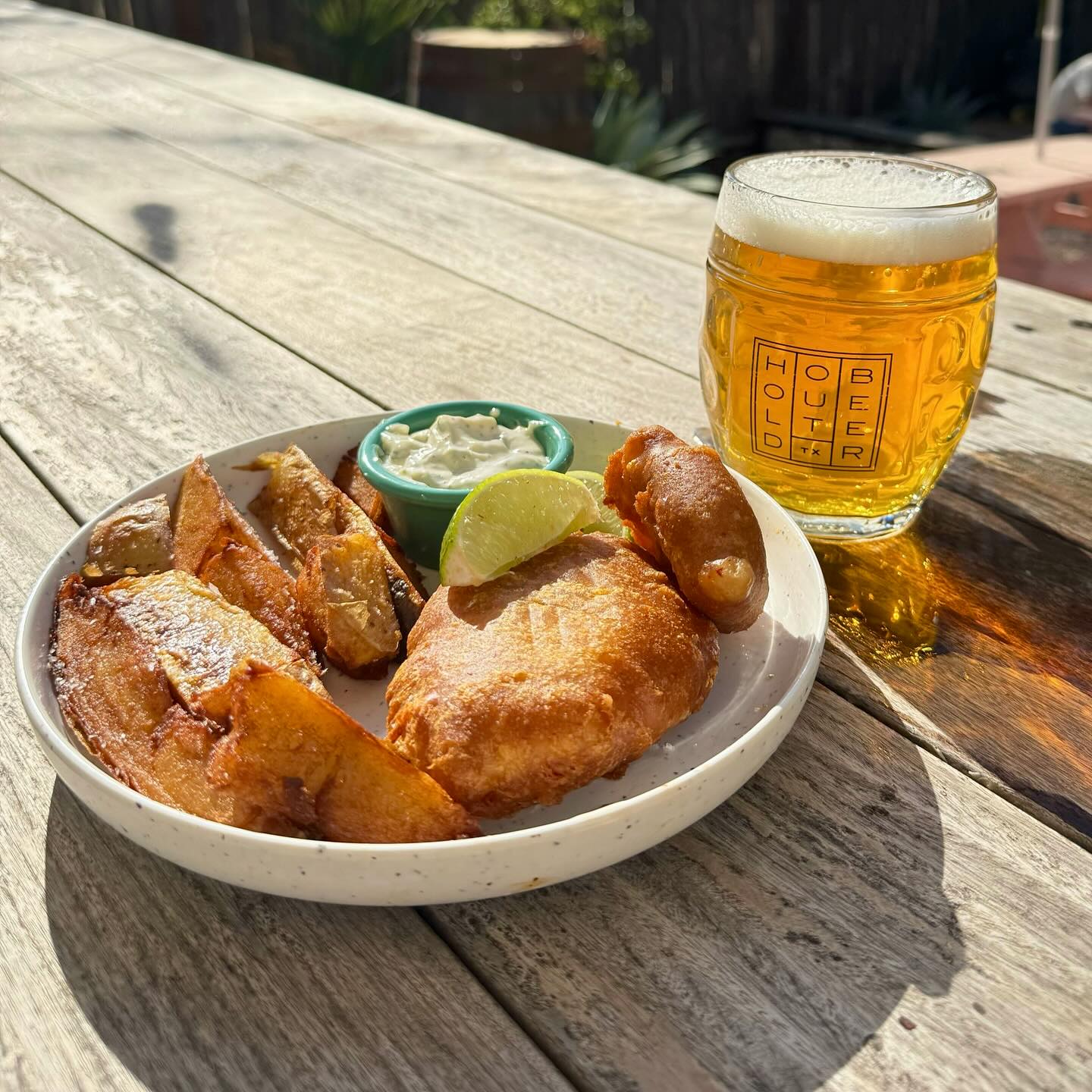 Fried fish, potato wedges, sauce, lime, and beer on an outdoor table.