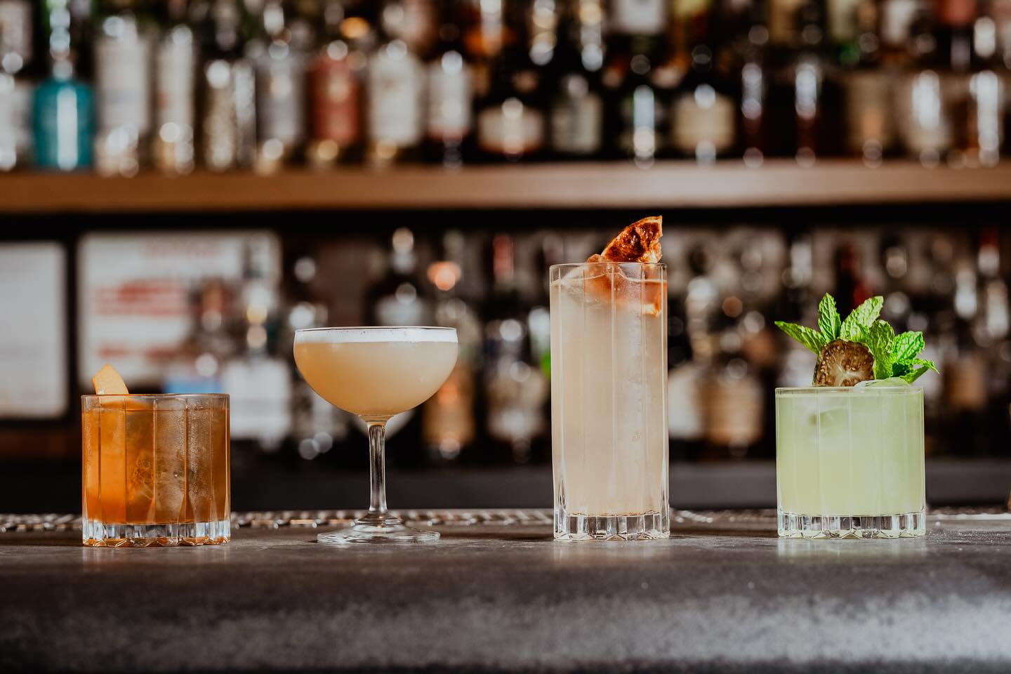 Four different cocktails on a bar counter with blurred bottles.