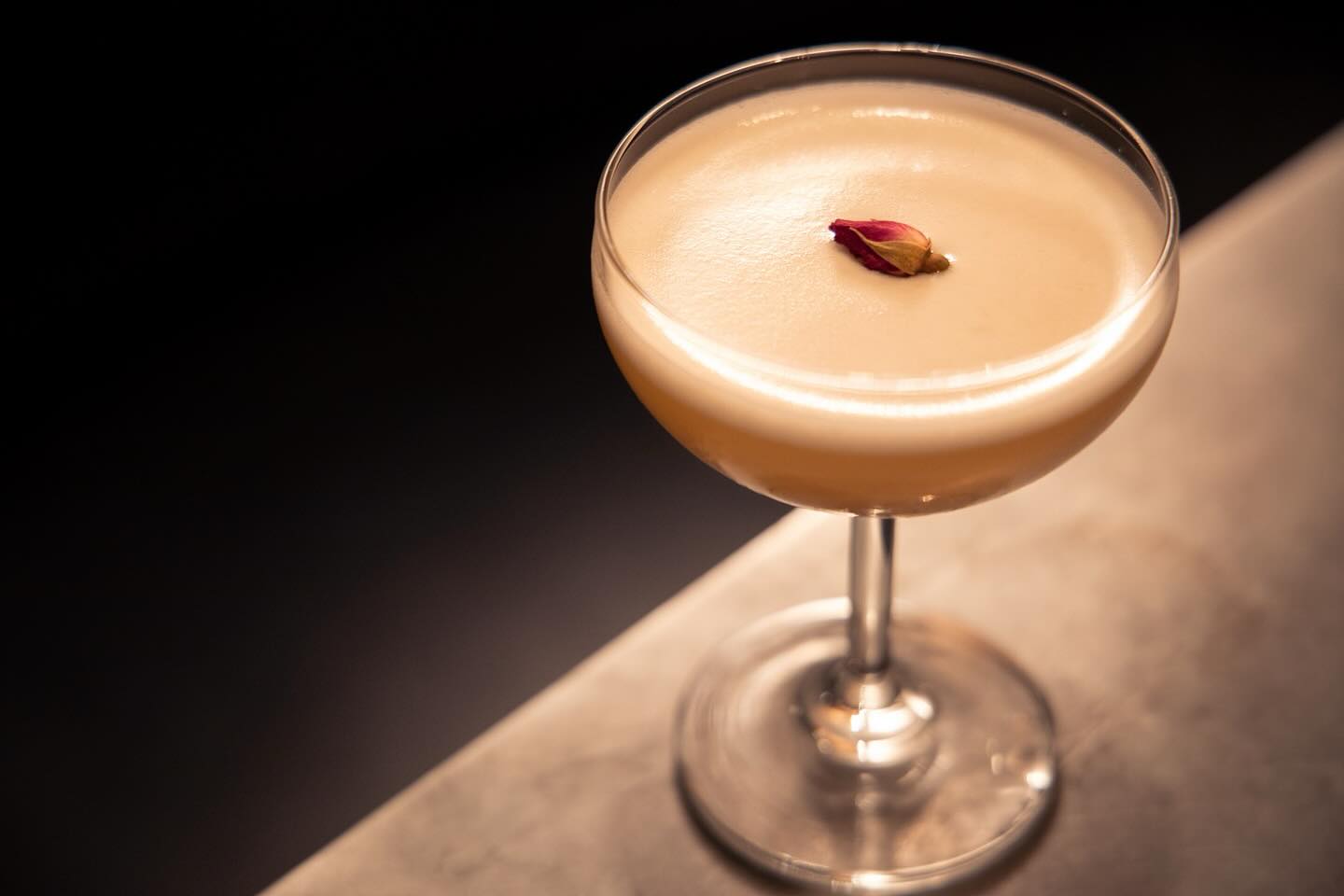 Foamy cocktail in a coupe glass garnished with a red rosebud.