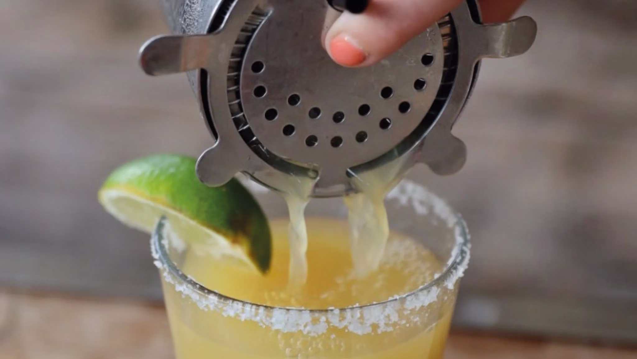 Hand straining yellow liquid into a salt-rimmed glass with a lime.