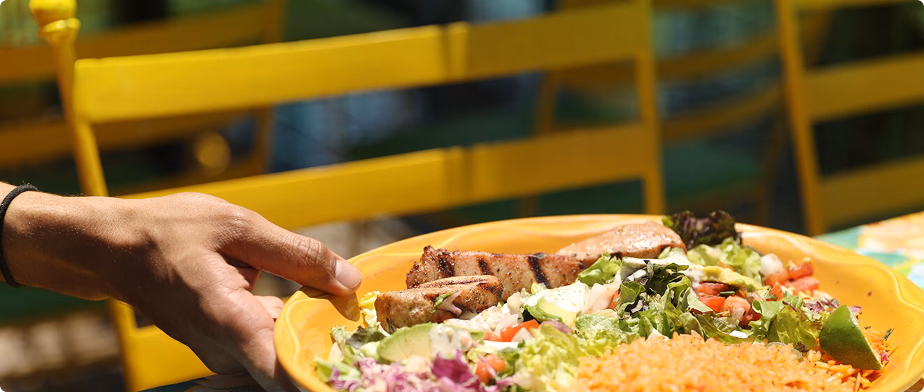 Hand holding a yellow bowl of grilled meat, salad, and rice.