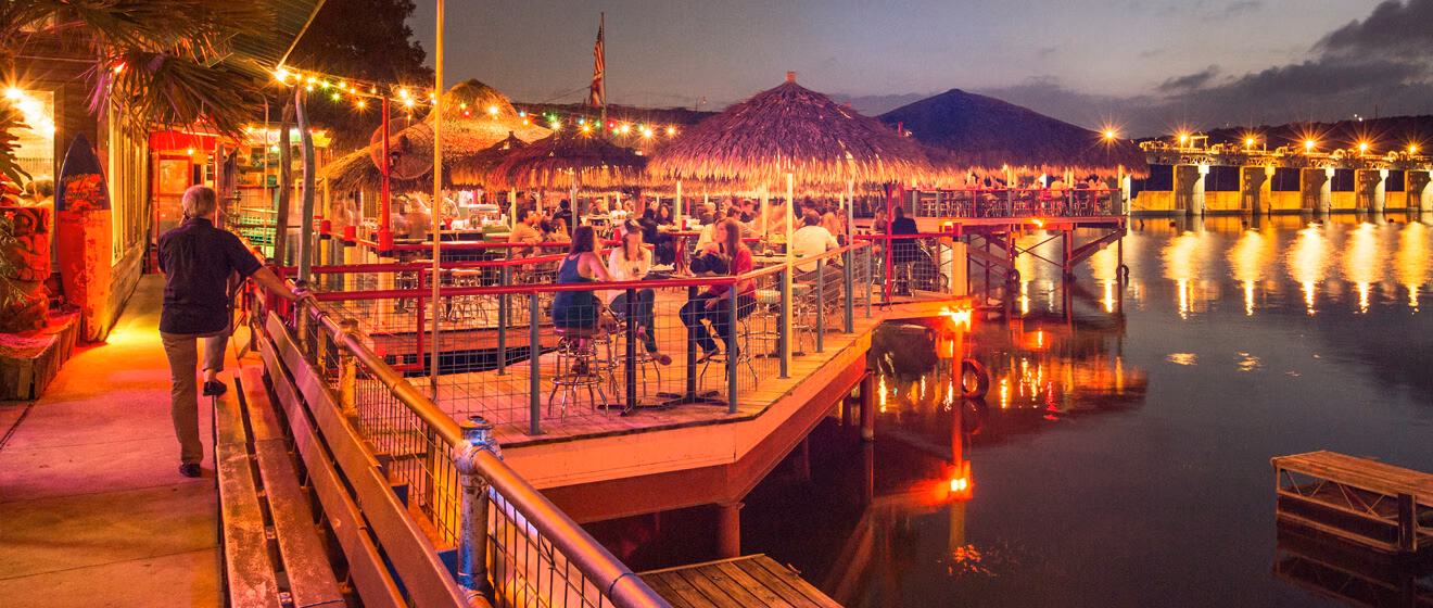 Illuminated outdoor restaurant with straw-roofed umbrellas by water at night.