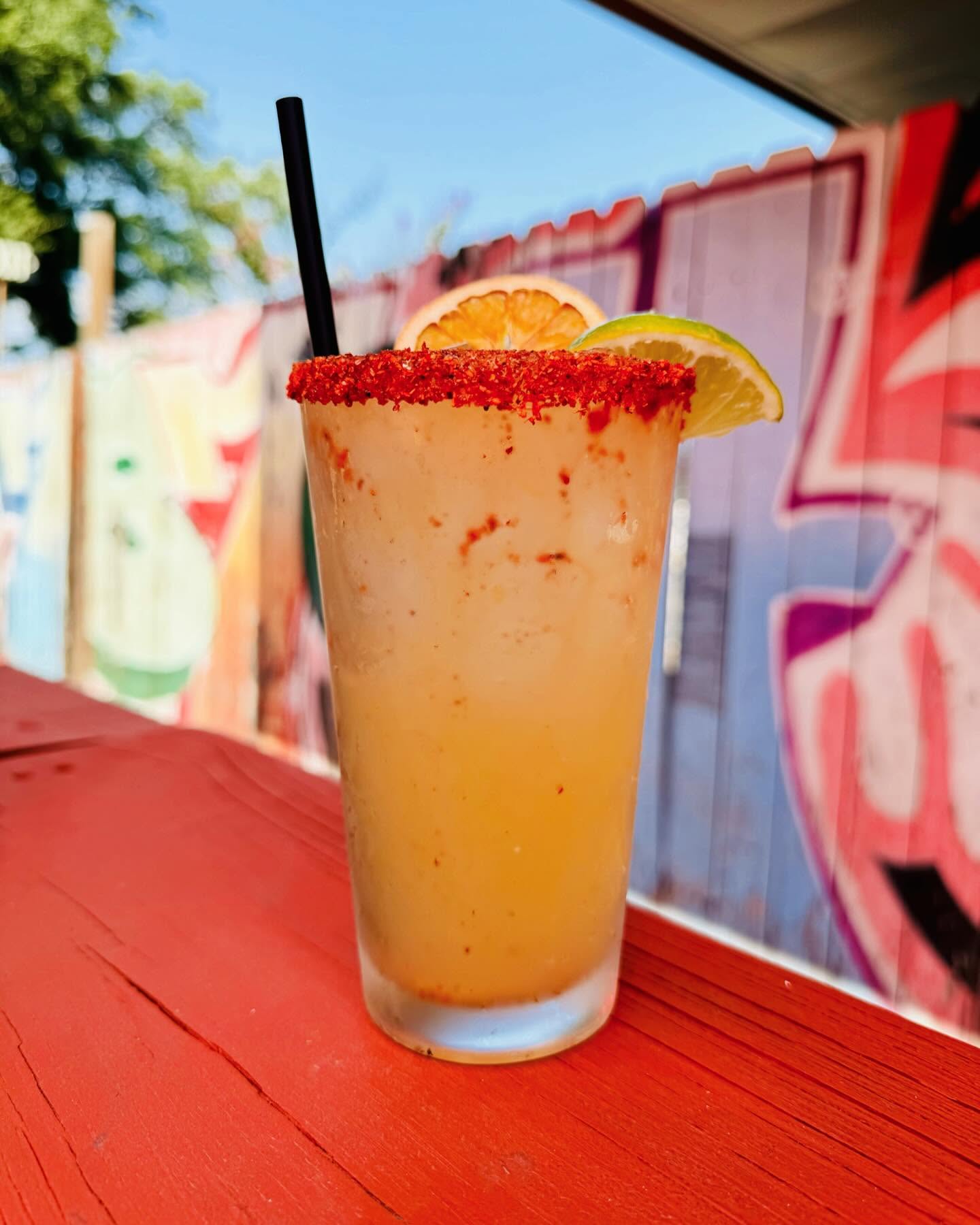 Frozen drink with chili salt rim, orange, and lime on red table.
