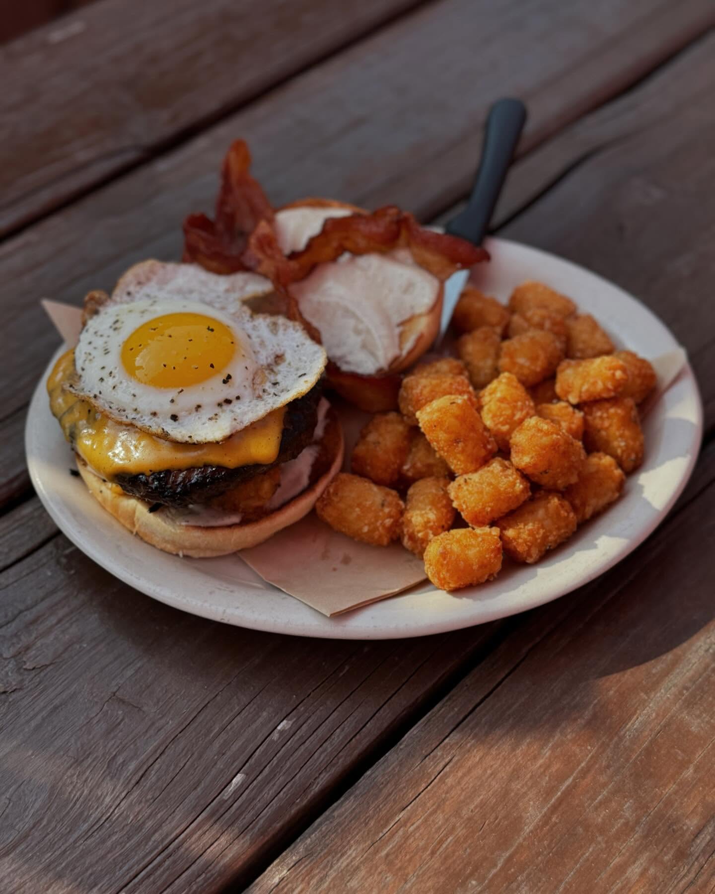 A bacon cheeseburger with a fried egg and tater tots on a wooden table.