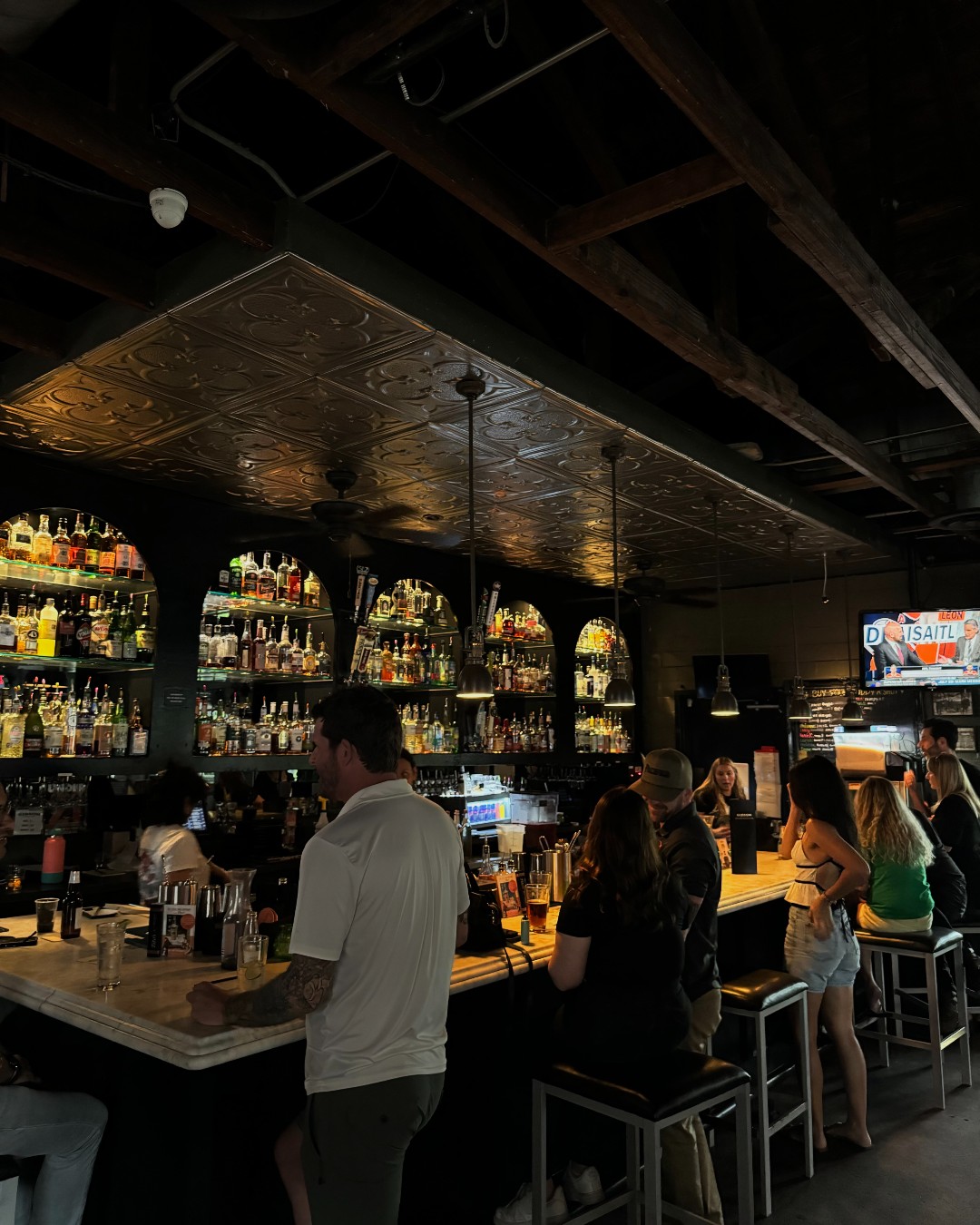 Dimly lit bar interior with patrons, liquor bottles, and tin ceiling.