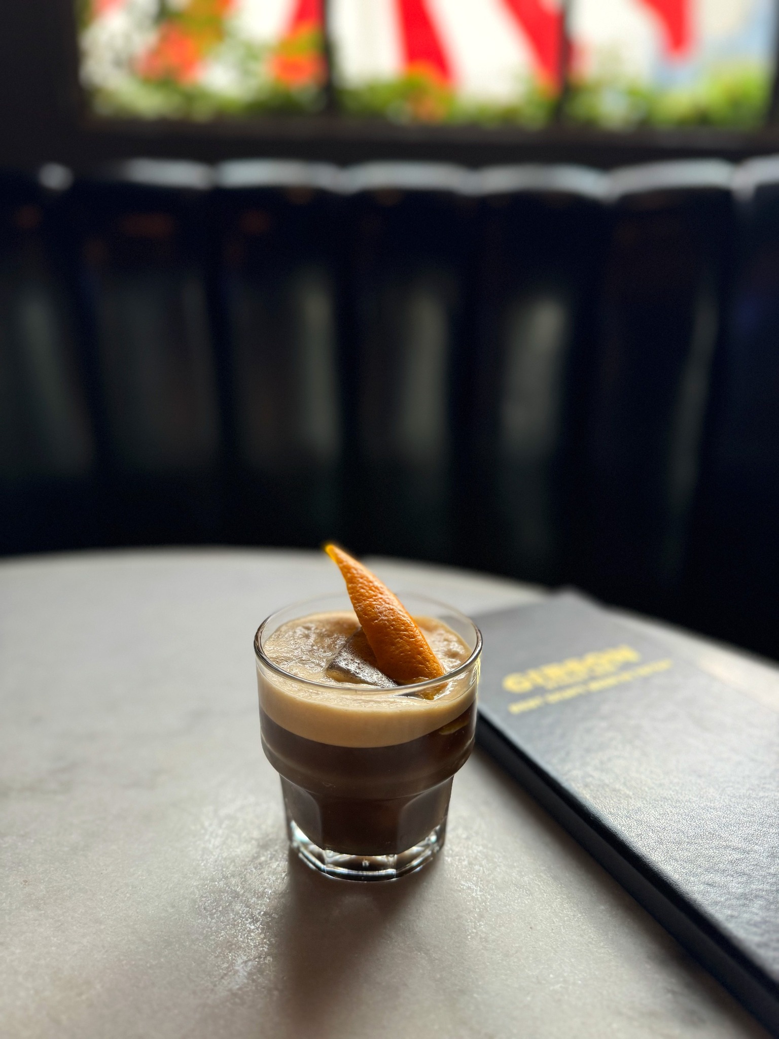 Dark layered drink with an orange peel and foam on a table.