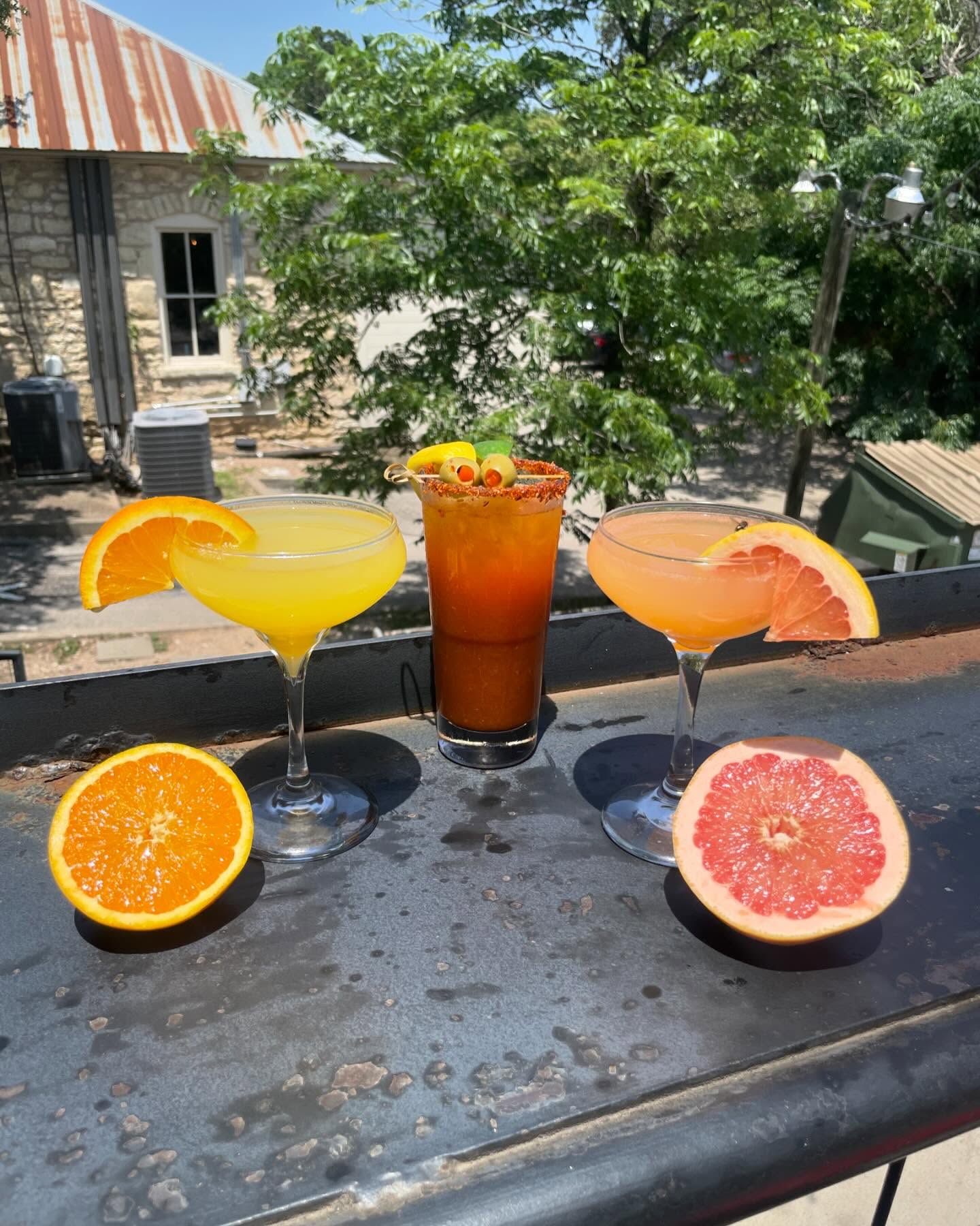 Three cocktails and citrus halves on an outdoor surface.