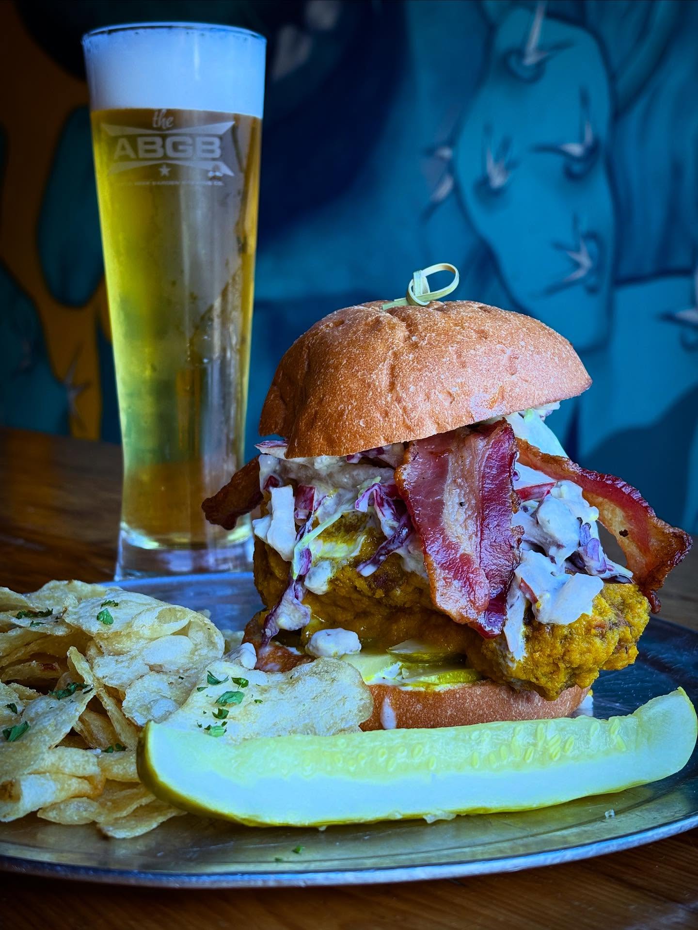 Fried chicken sandwich with bacon, coleslaw, chips, pickle, and beer.