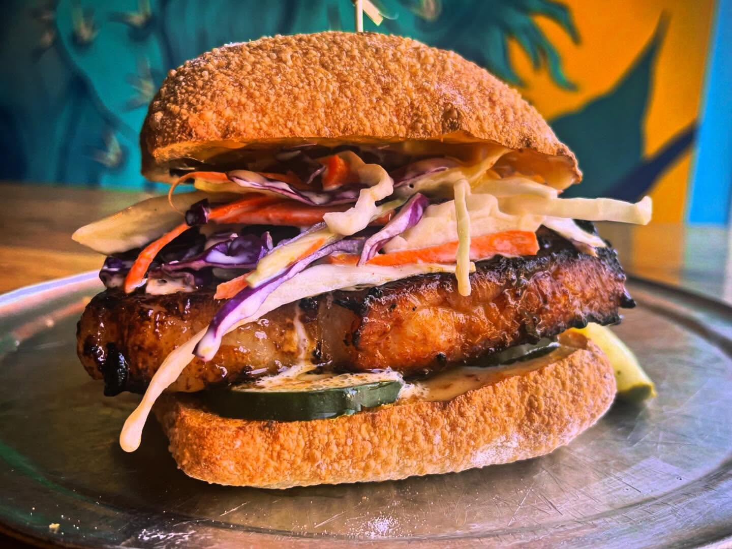 Burger with glazed meat, colorful coleslaw, and pickles on a bun.