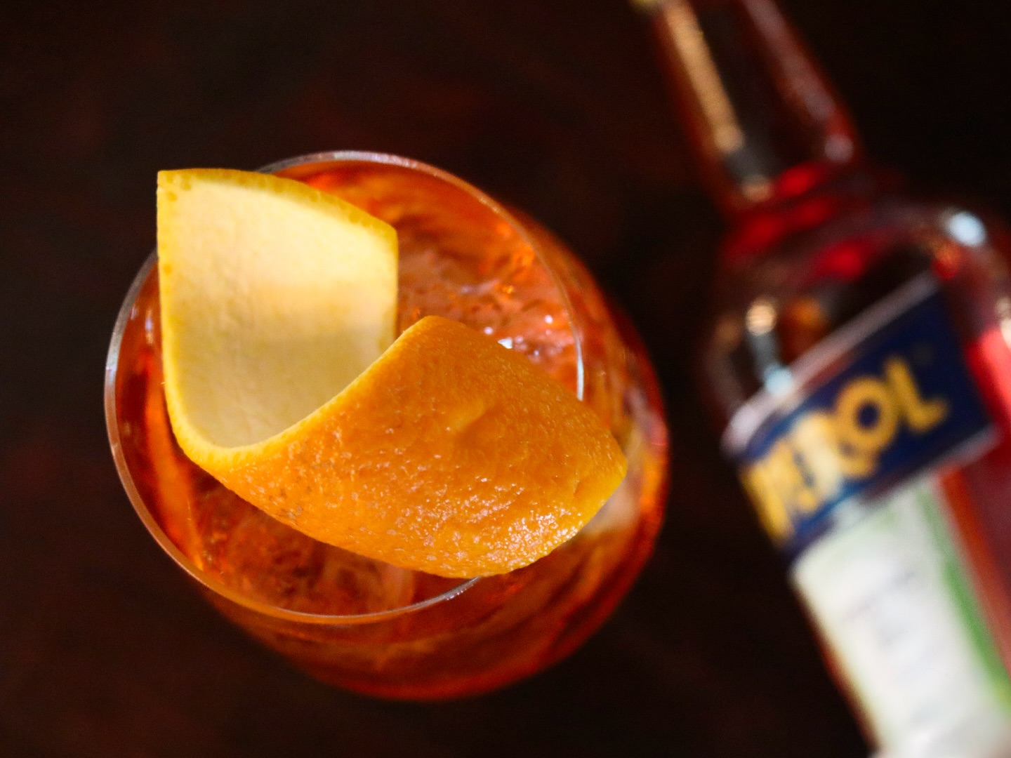 Orange-colored drink with an orange peel and a blurred Aperol bottle.
