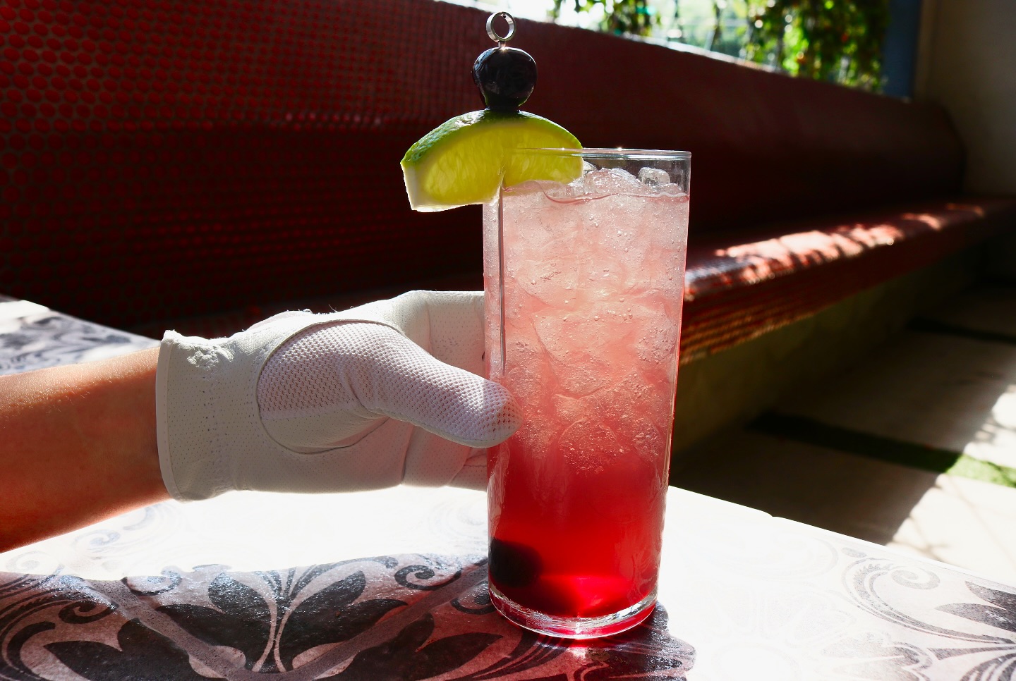 Hand in white glove holds tall iced pink drink with lime.