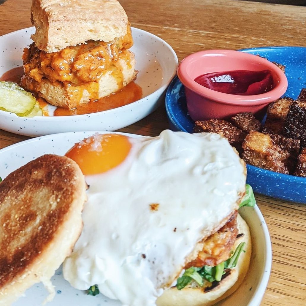 Fried chicken biscuit, egg sandwich, and potato cubes on a wooden table.