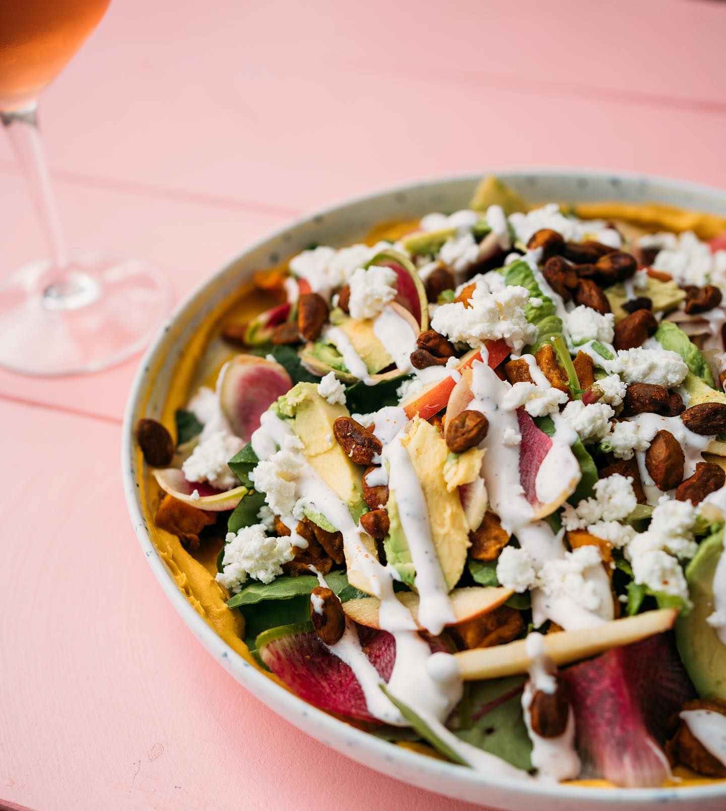 Salad with greens, avocado, radish, crumbled cheese, and dressing on a pink table.