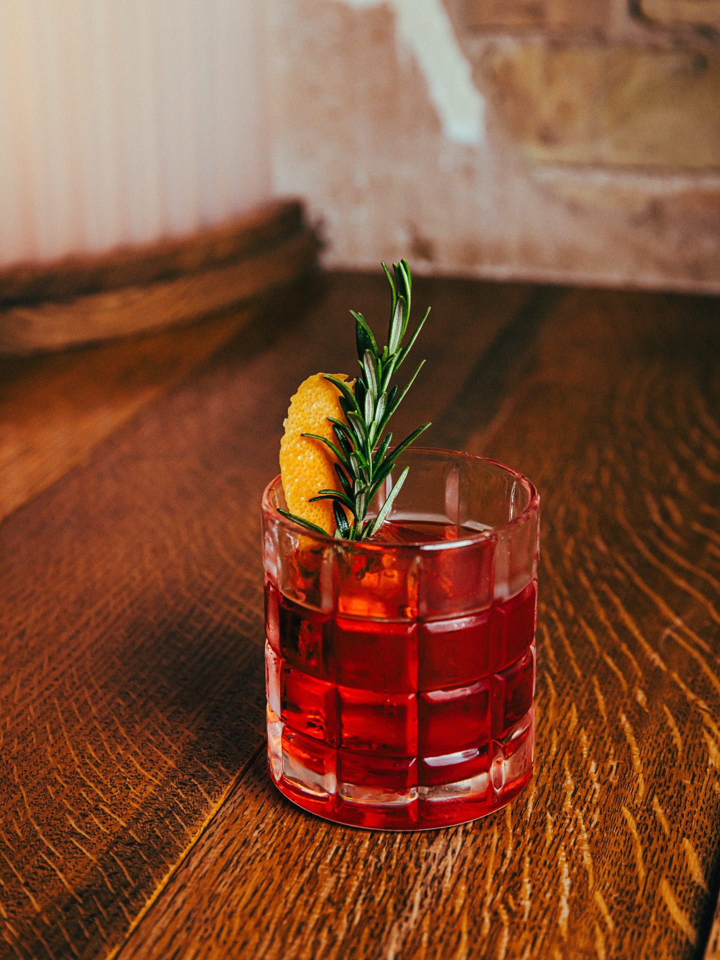 Red cocktail with orange peel and rosemary on a wooden table.