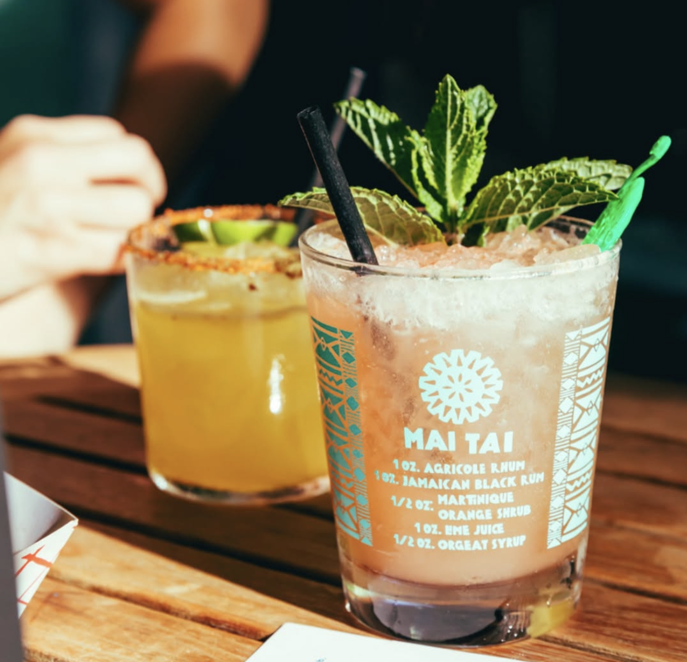 Two cocktails on a wooden table, one a Mai Tai with mint.