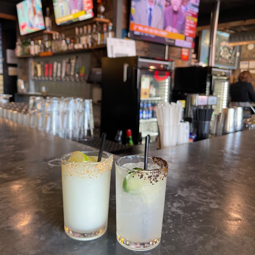 Two different cocktails with lime wedges on a bar.