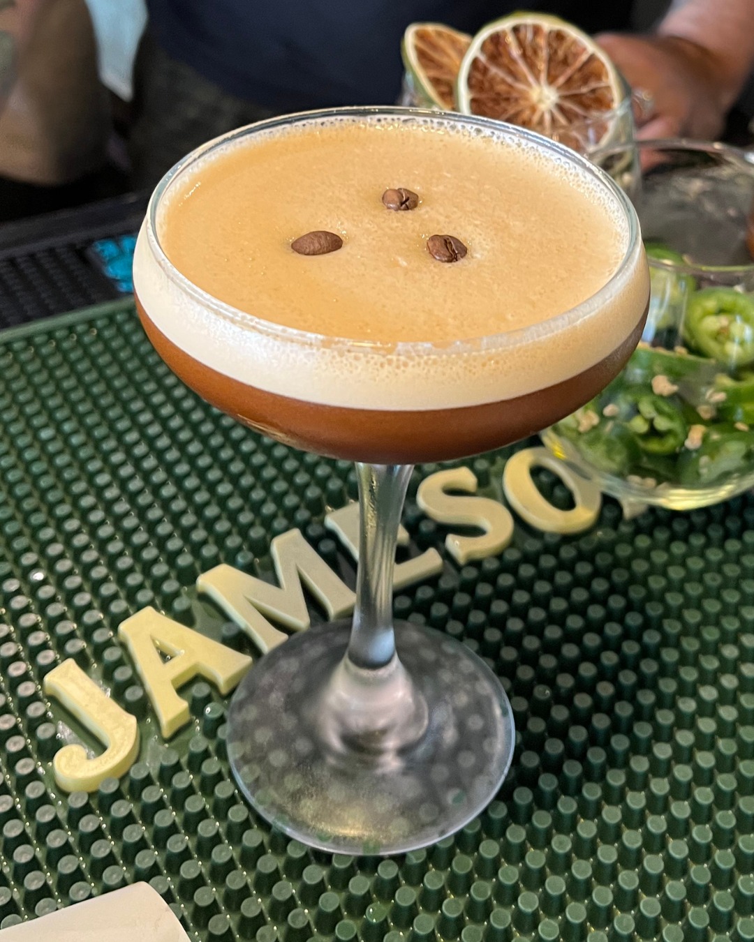Cocktail with frothy top, coffee beans, and JAMESON letters on mat.