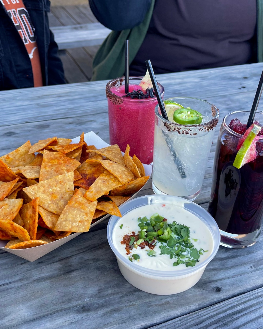Chips, queso dip, and three beverages on a rustic wooden table.