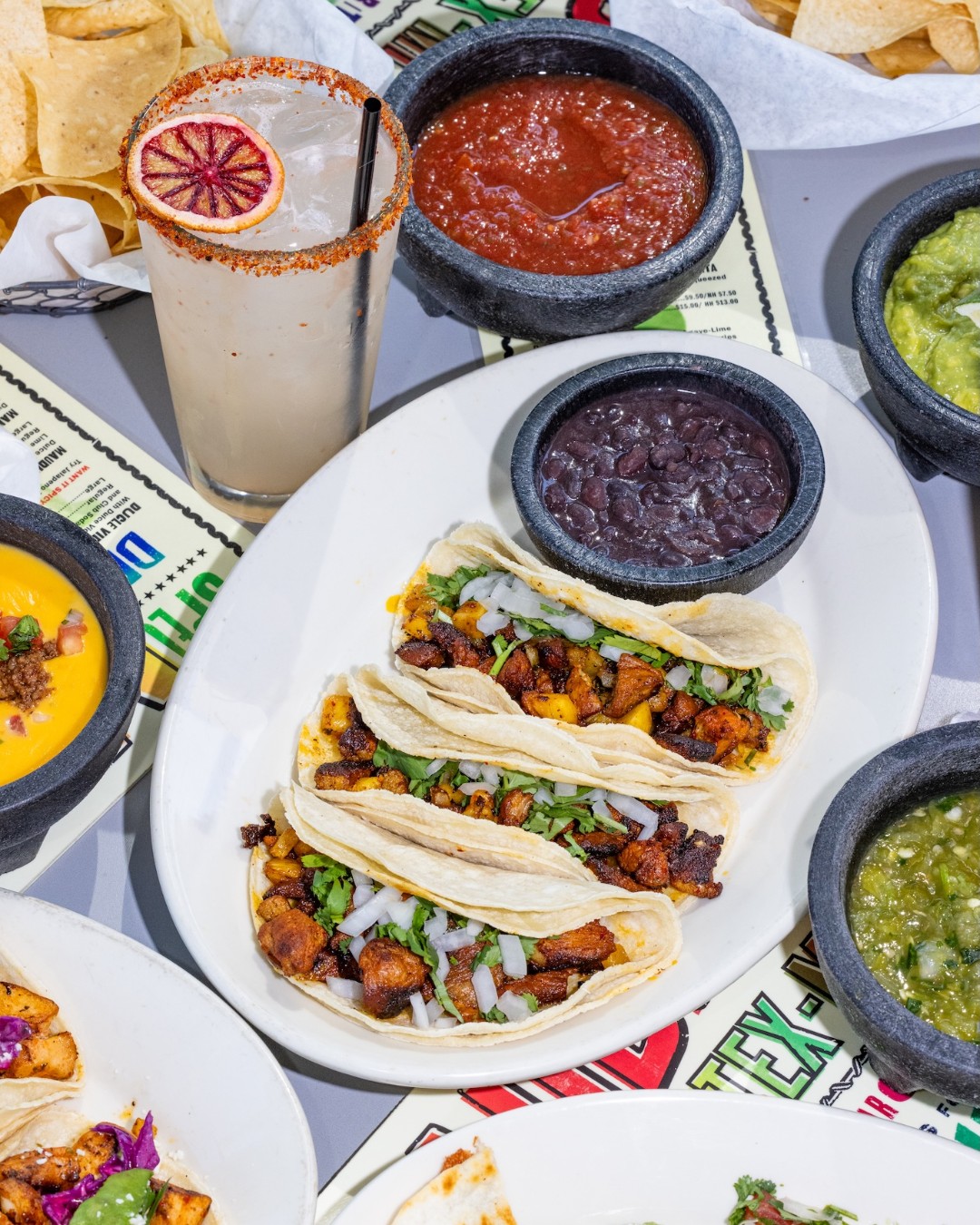 Tacos, chips, salsa, guacamole, queso, black beans, and a cocktail.