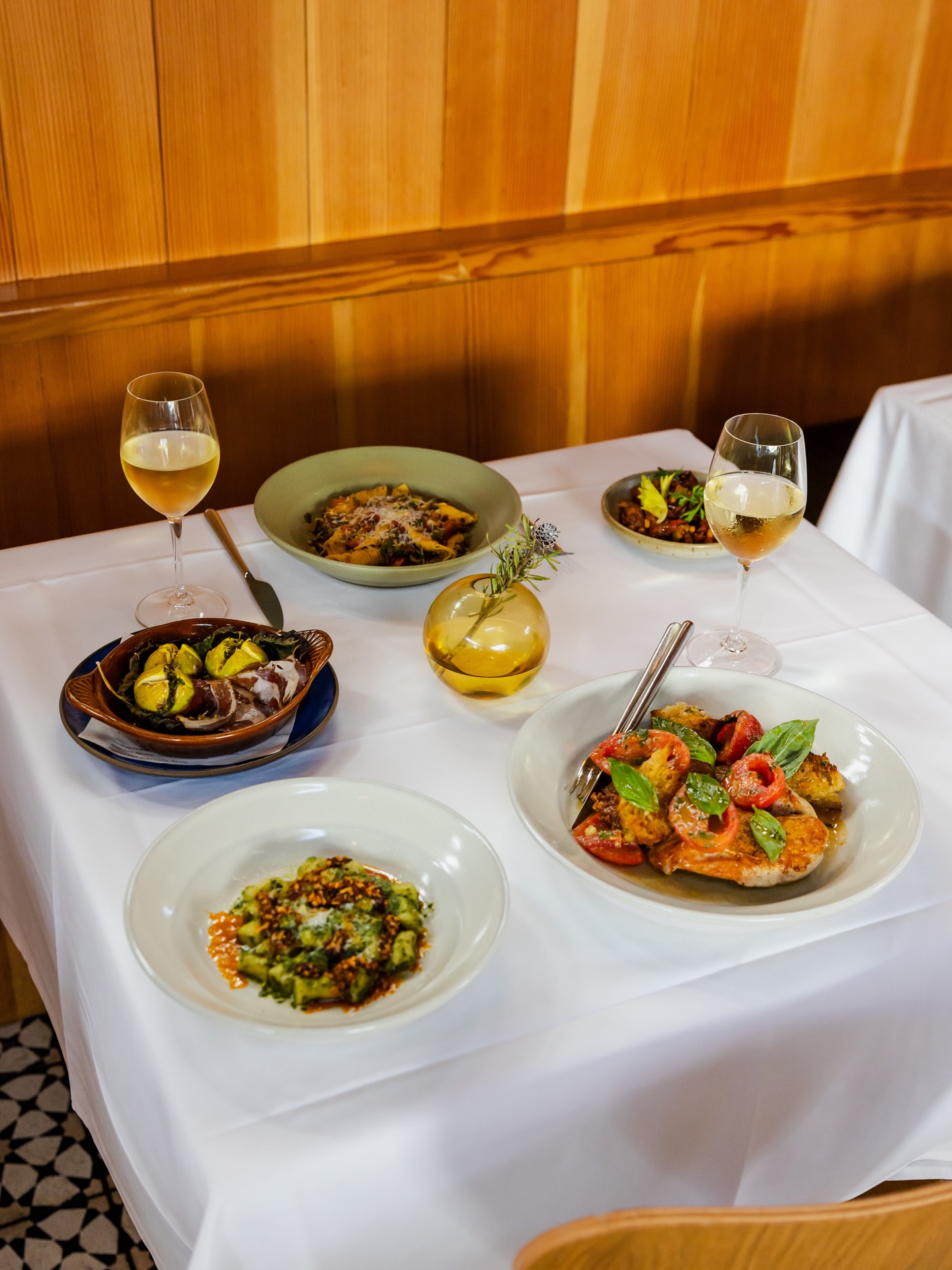 Dining table with multiple plates of food, white wine, and wooden wall.