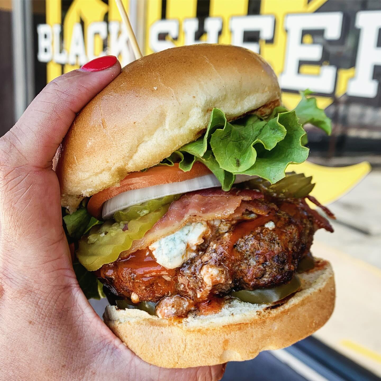 Hand holding a burger with lettuce, tomato, onion, pickles, bacon, blue cheese.