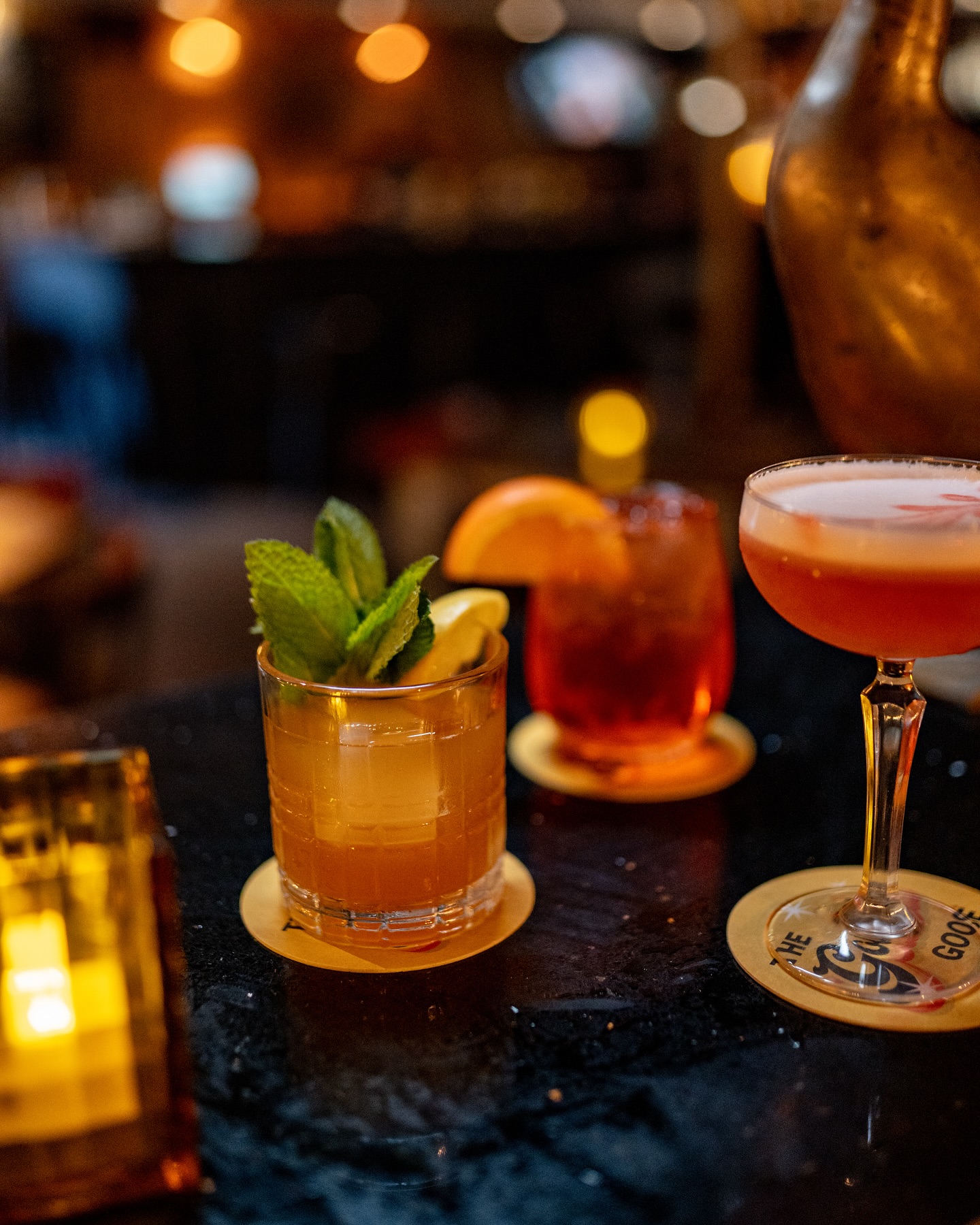 Three cocktails on a dark bar, with blurred background lights.