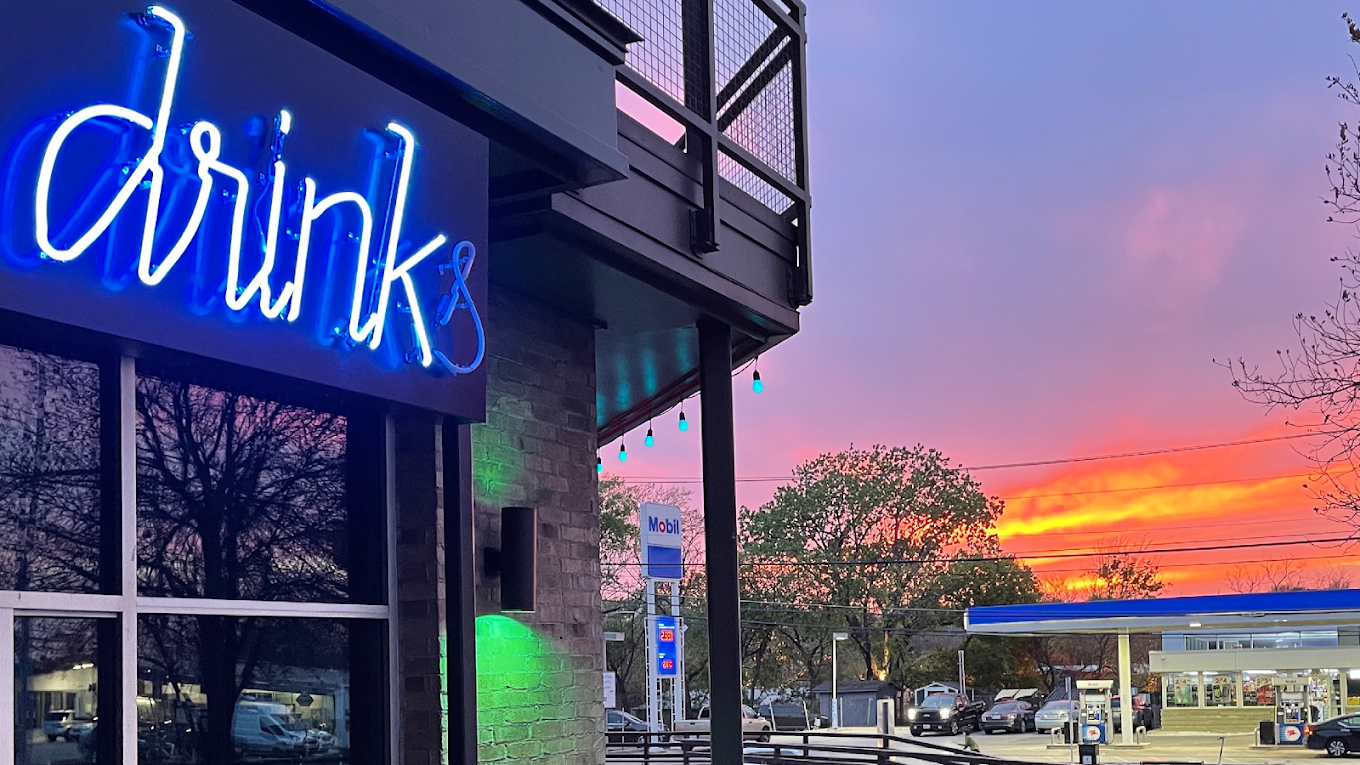 Blue neon "drinks" sign, gas station, and colorful sunset at dusk.