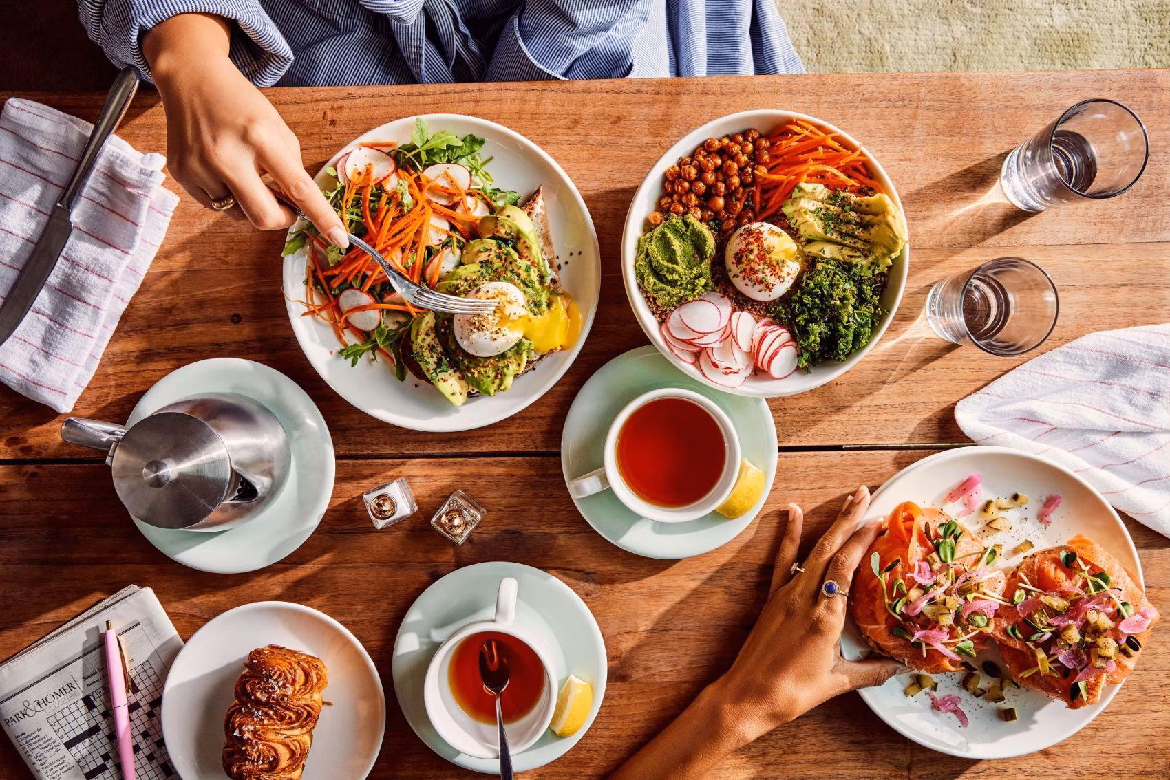 Hands reach for diverse breakfast dishes and drinks on a wooden table.