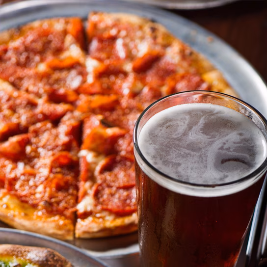 Glass of dark beer with blurred pepperoni pizza in background.