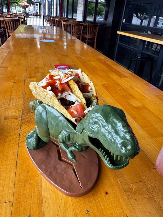 Two tacos in a green dinosaur holder on a wooden table.