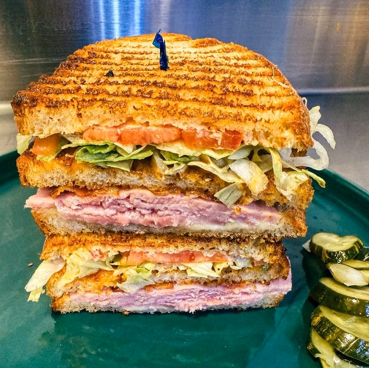Stacked grilled ham sandwich with lettuce, tomato, and pickles.