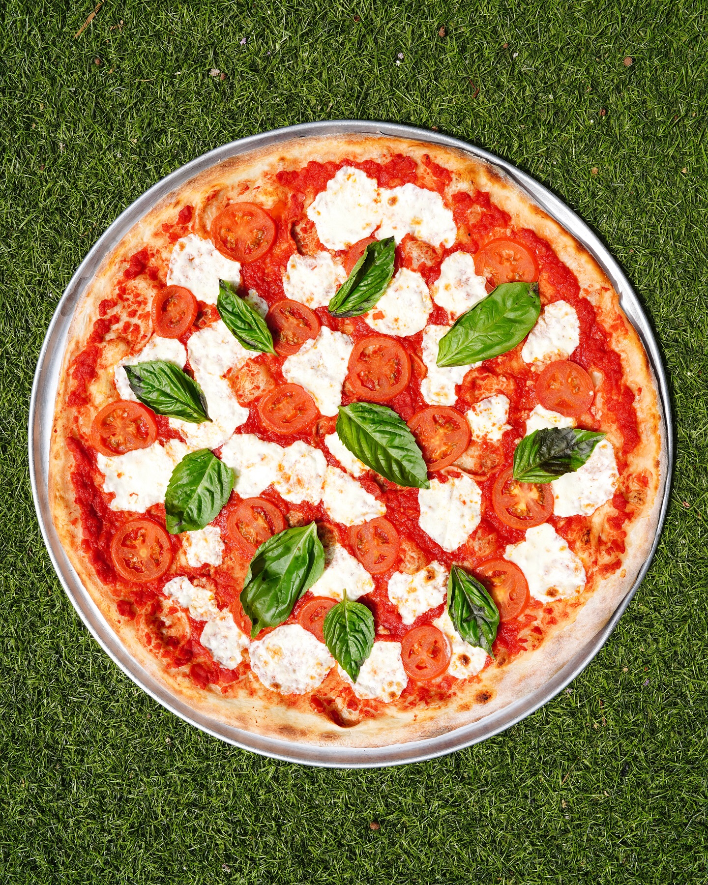 Margherita pizza with mozzarella, tomatoes, and basil on green grass.