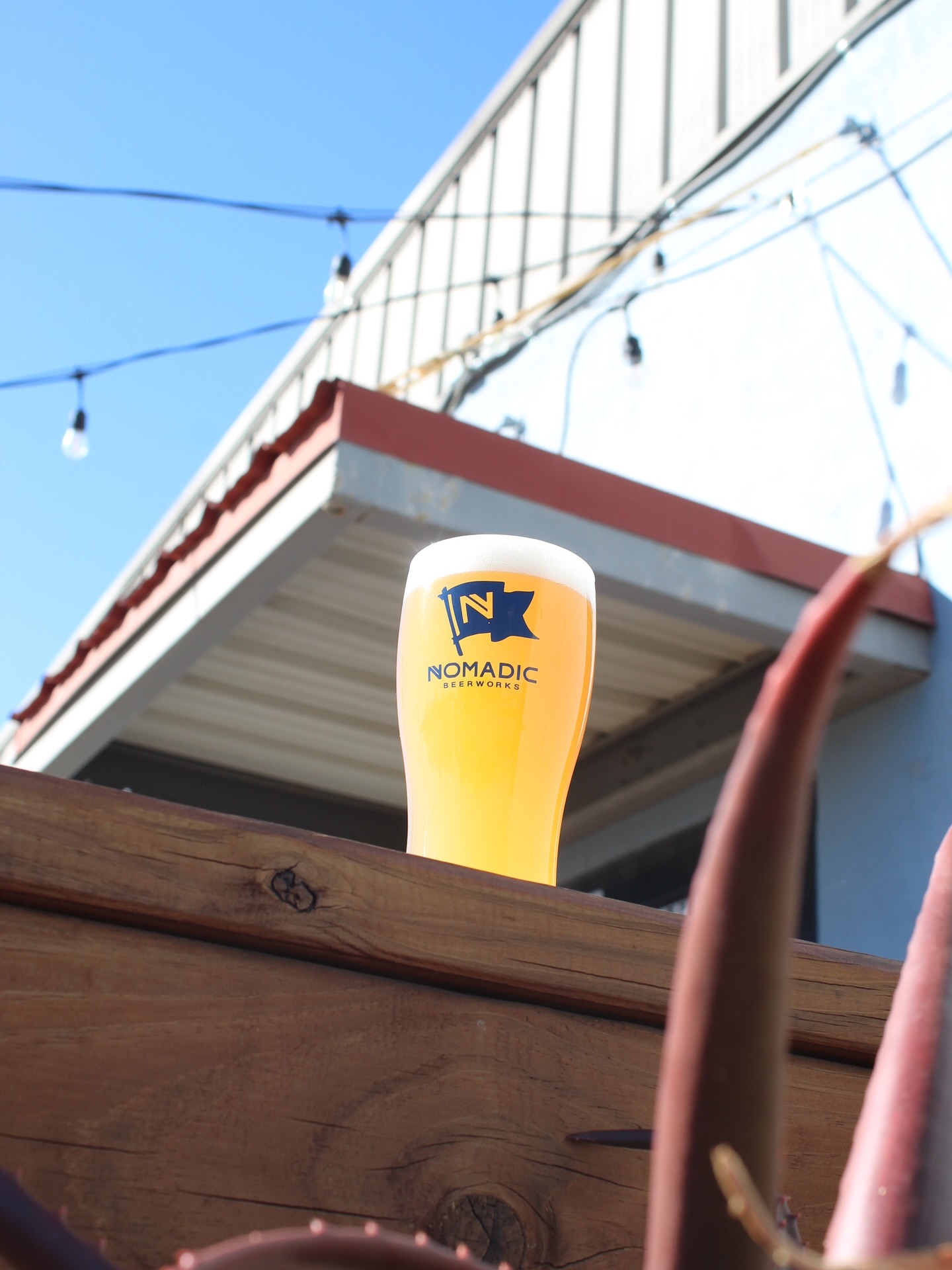 Glass of Nomadic Beerworks beer on a wooden railing outdoors.