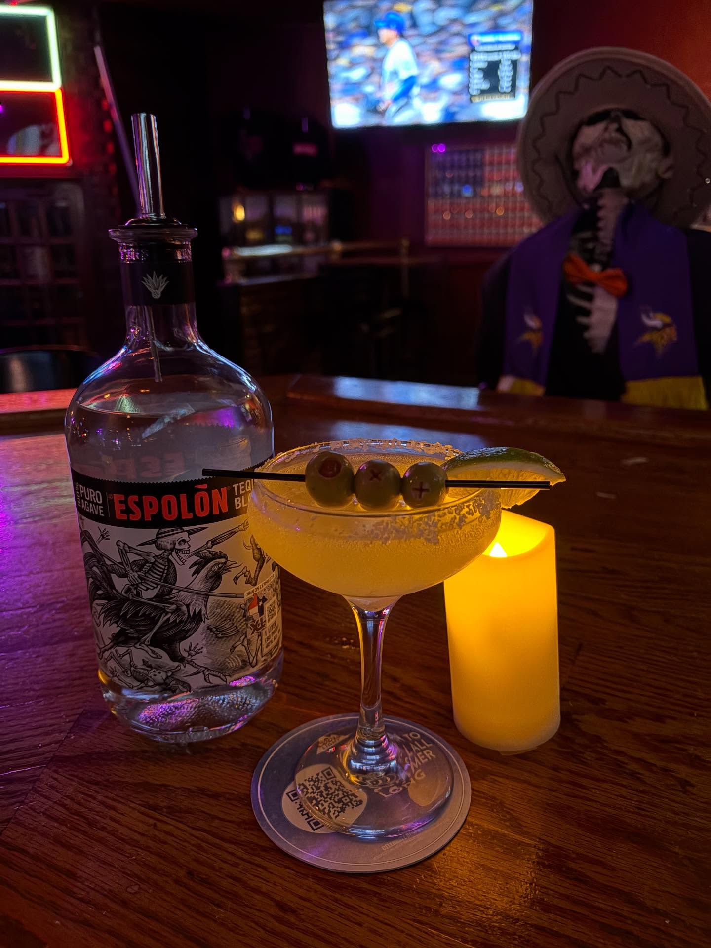 Espolon tequila, olive-garnished cocktail, candle, skeleton at bar.