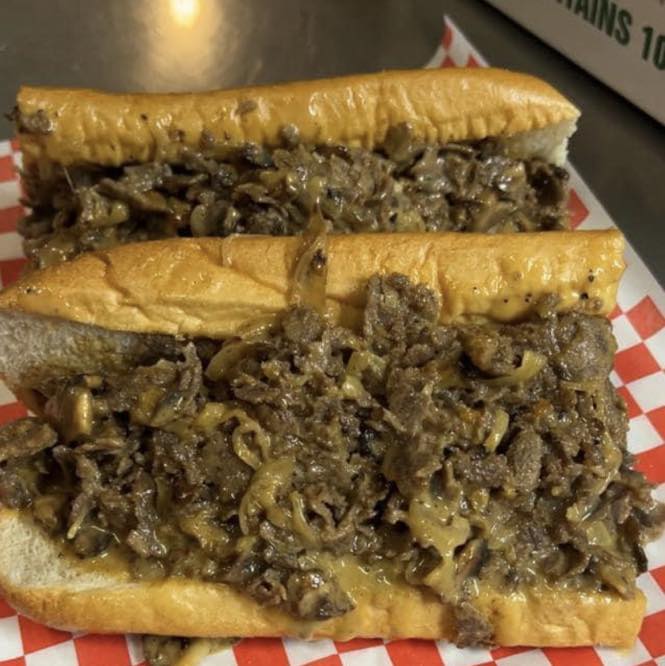 Two cheesesteak sandwiches on red and white checkered paper.