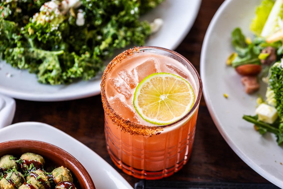 Pink cocktail with lime slice and spiced rim, surrounded by food.