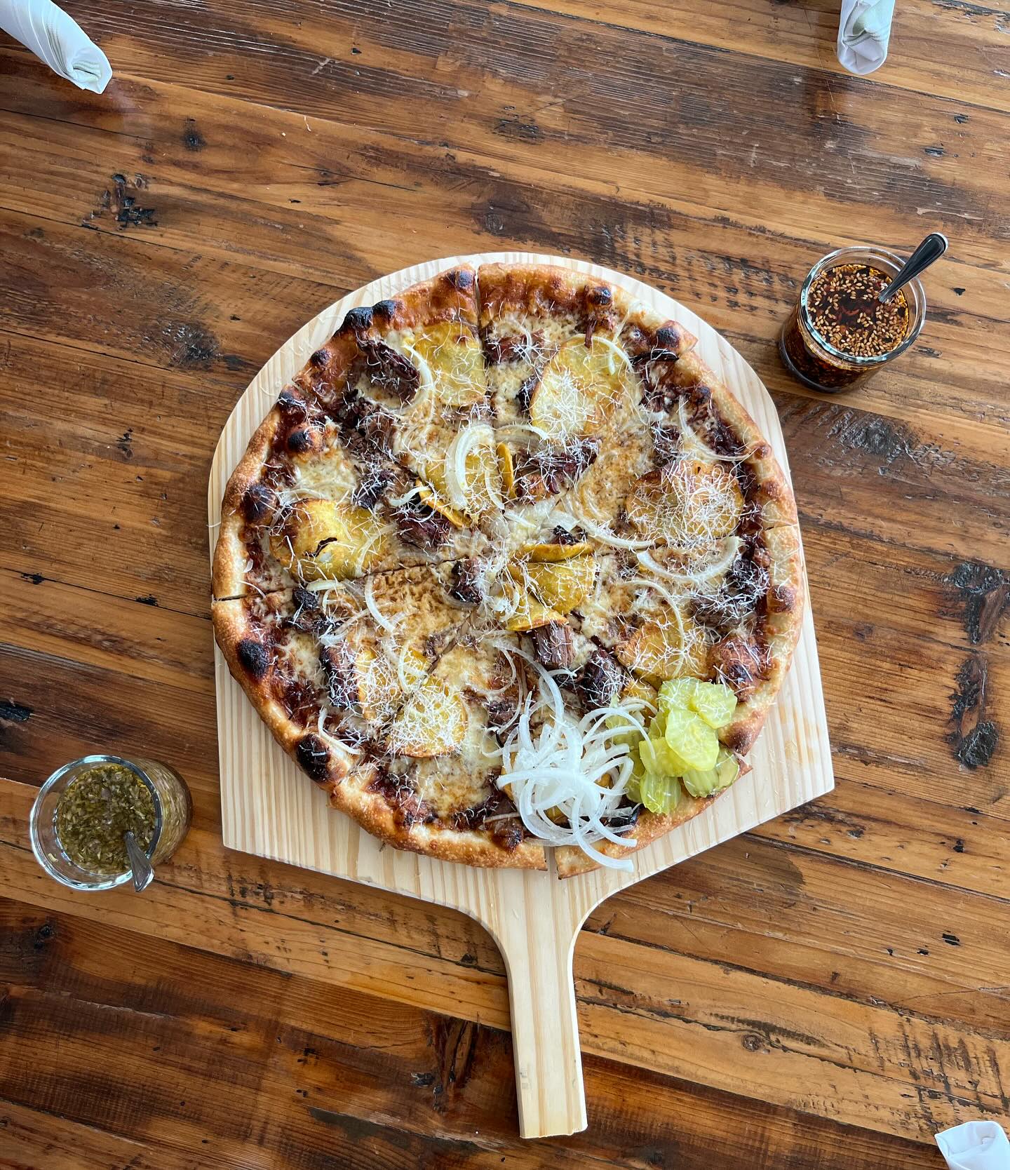Pizza topped with potato slices, onions, cheese, and pickles on a peel.