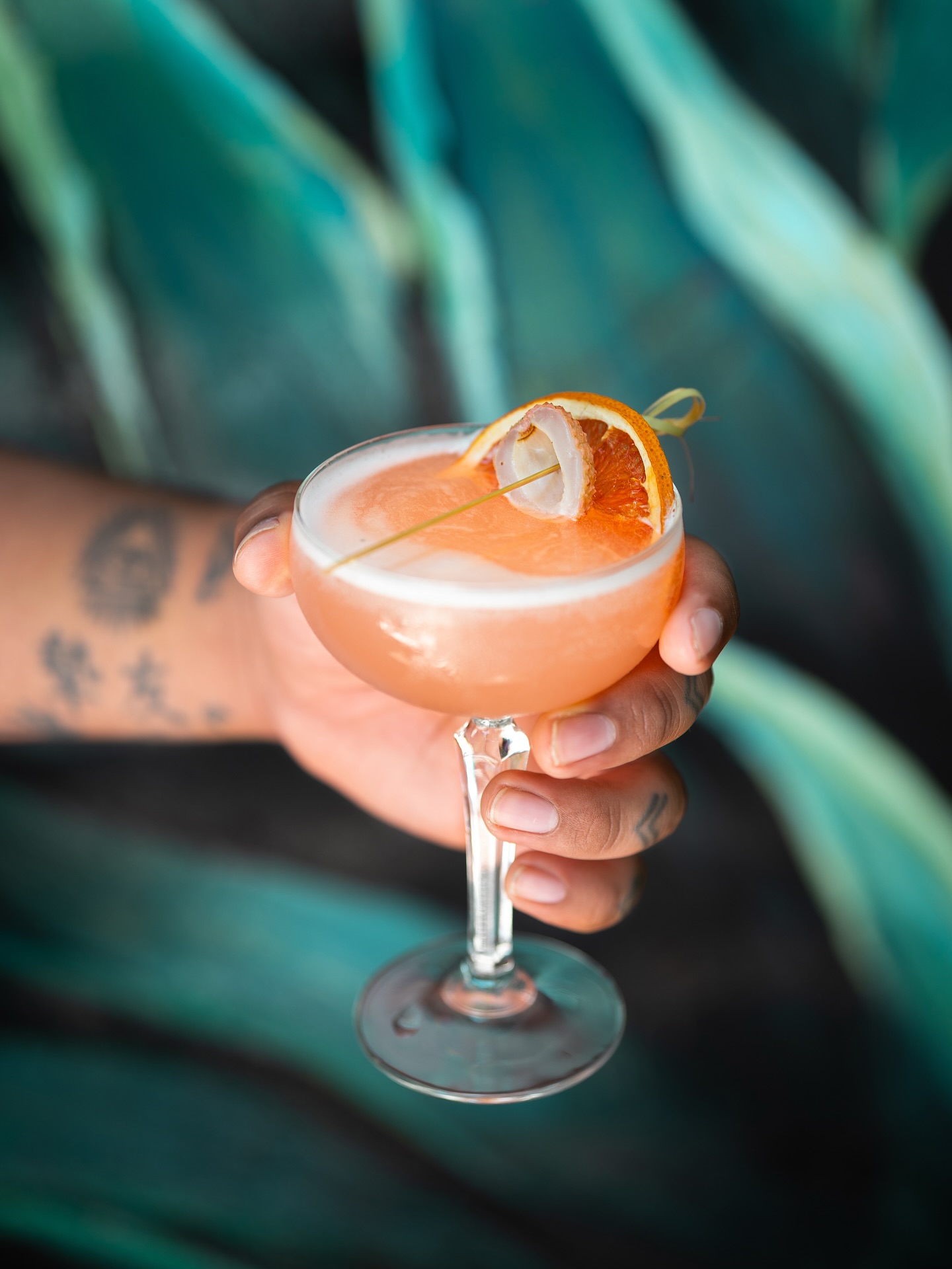 Tattooed hand holding a foamy light orange cocktail with orange garnish.