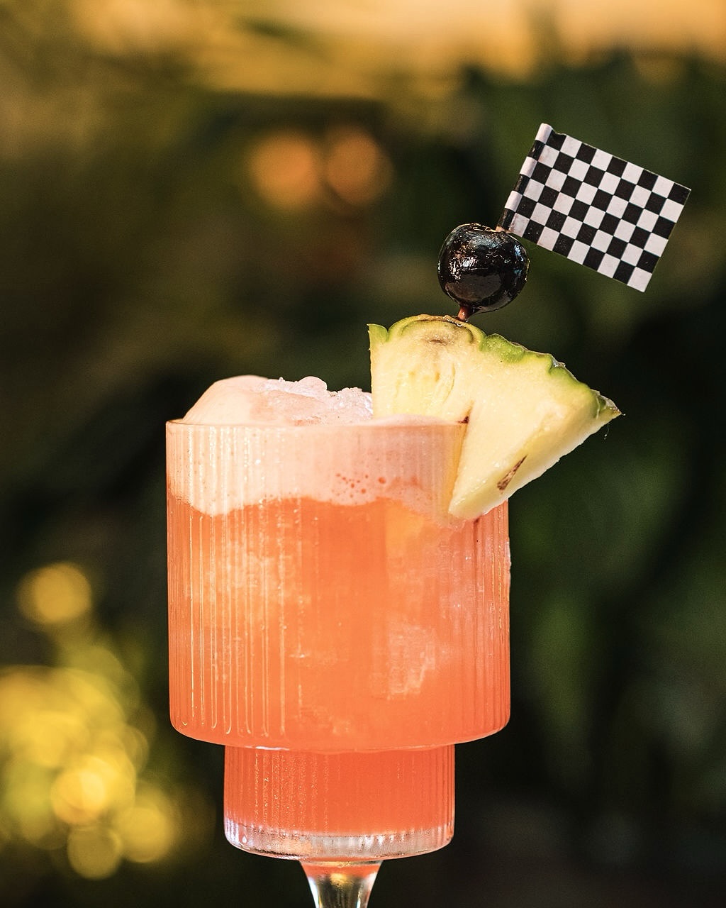 Orange cocktail in ribbed glass, garnished with pineapple, cherry, and checkered flag.
