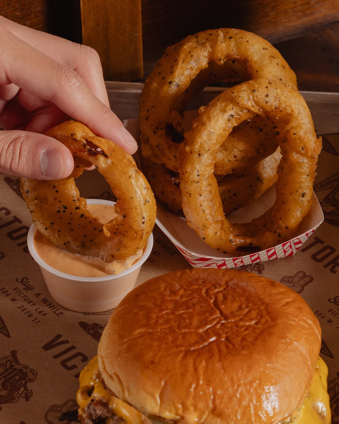 A hand dips a seasoned onion ring into sauce next to a cheeseburger.