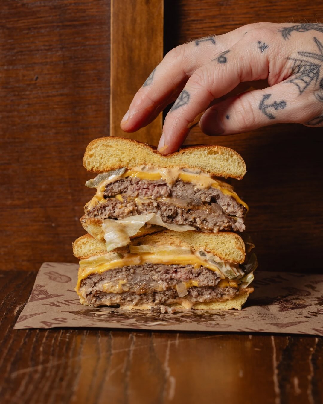 Tattooed hand presses a stacked burger with multiple patties and cheese.