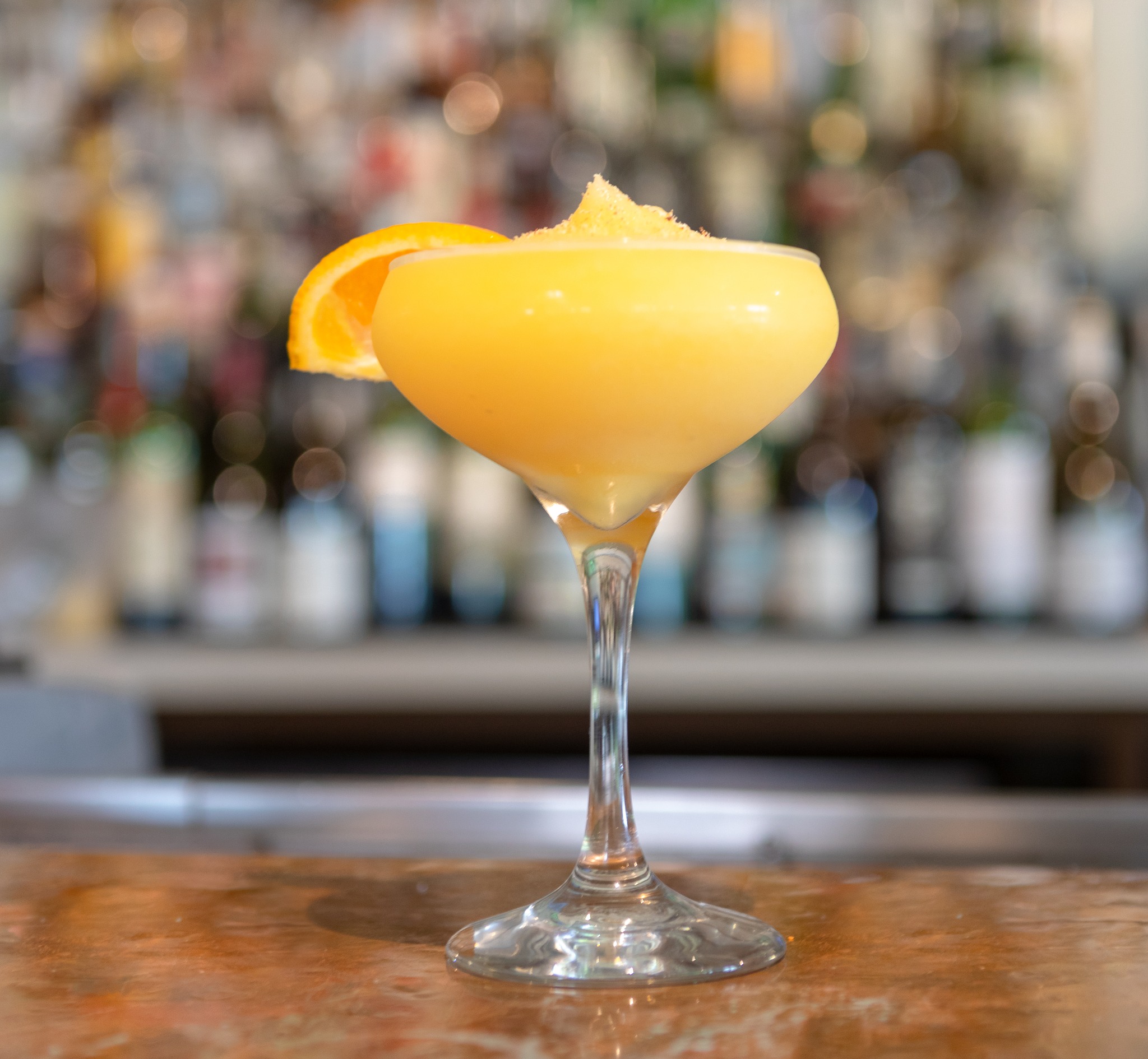 Yellow cocktail in stemmed glass with orange slice and granular topping.