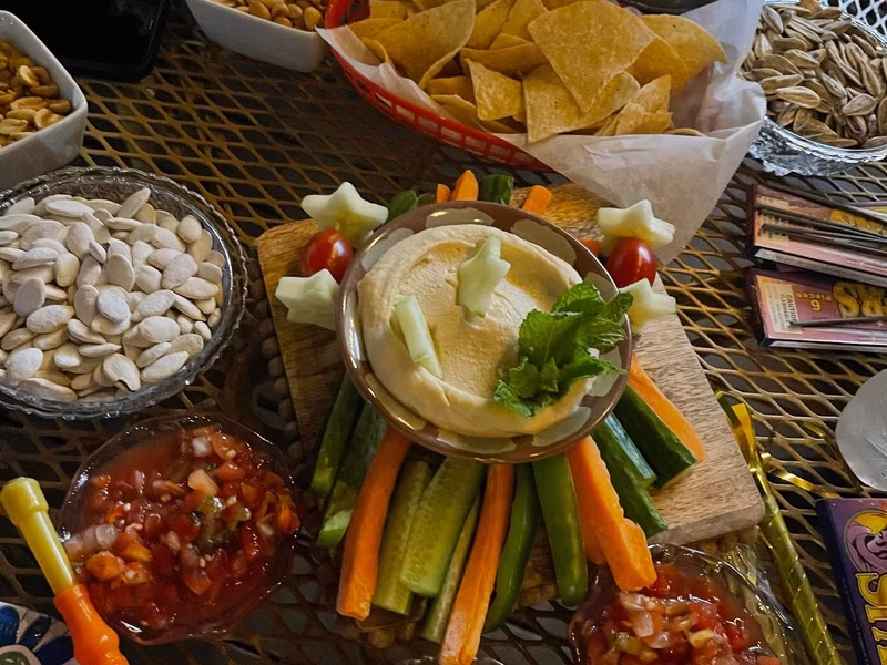 Hummus dip surrounded by vegetables, with chips, salsa, and seeds.
