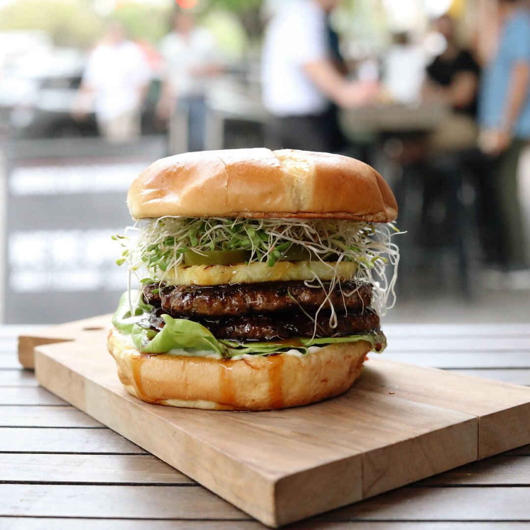 Large burger with two patties, grilled pineapple, sprouts, on a wooden board.