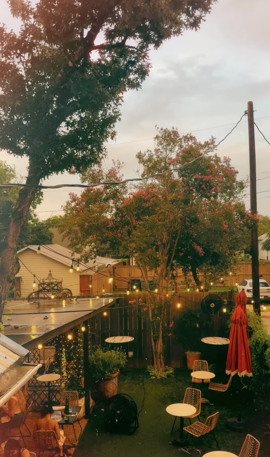 Outdoor patio with string lights, tables, chairs, people, and trees.