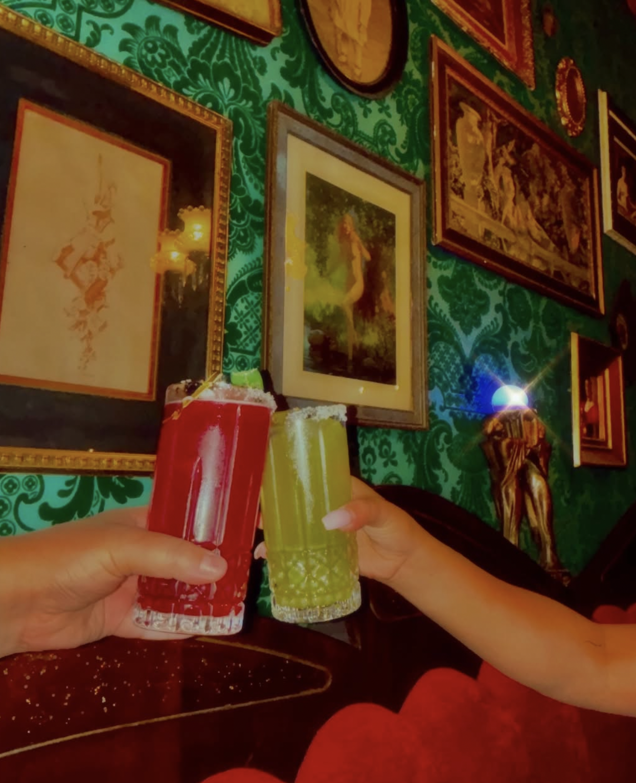 Two hands clinking red and yellow cocktails against an ornate green wall.