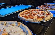 Three large pizzas on tables with blue pool tables in background.
