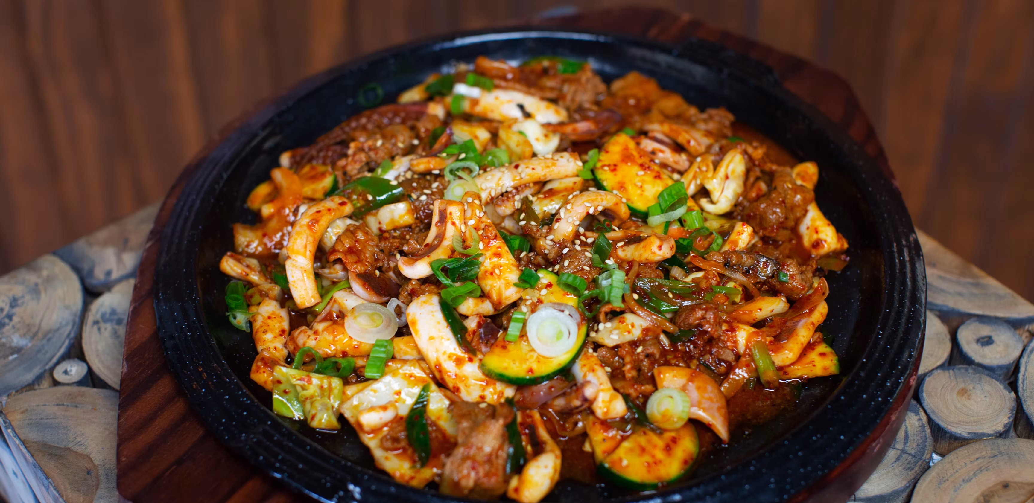 Spicy dish with squid, meat, zucchini, and scallions in a dark pan.