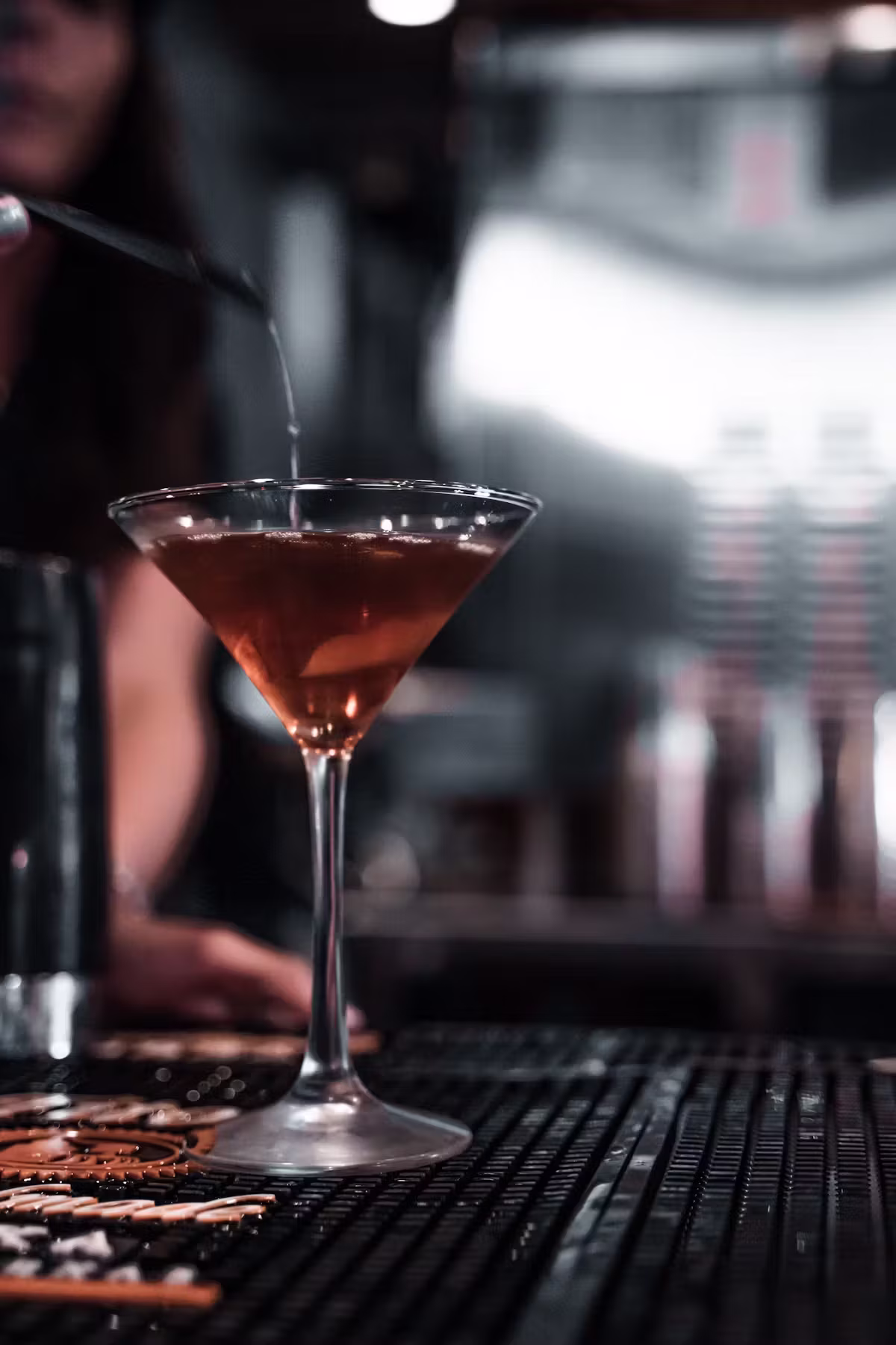Bartender pouring liquid into a reddish cocktail in a martini glass.